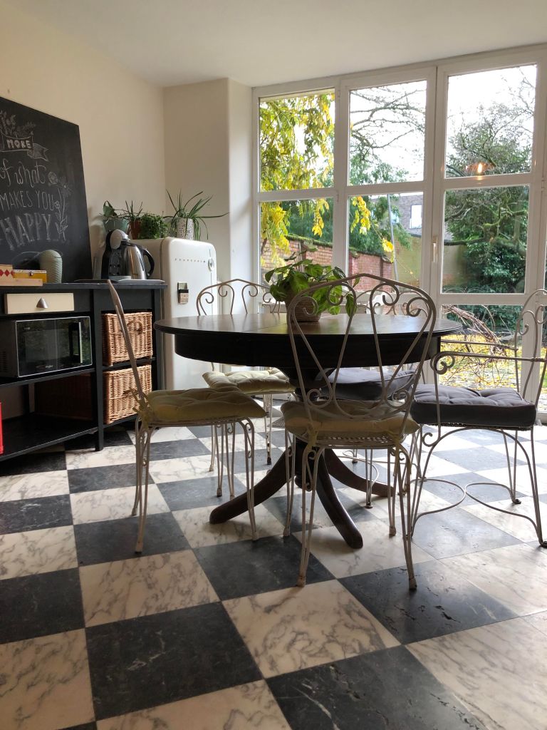 Gezellige keuken met een ronde tafel, metalen stoelen en grote ramen met uitzicht op de tuin. De vloer is afgewerkt met tegels in zwart-wit patroon.