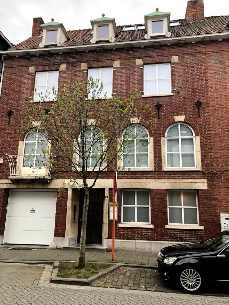 Gevel van een bakstenen appartement met gedeelde ingang, een kleine boom vooraan en een zwarte auto geparkeerd.